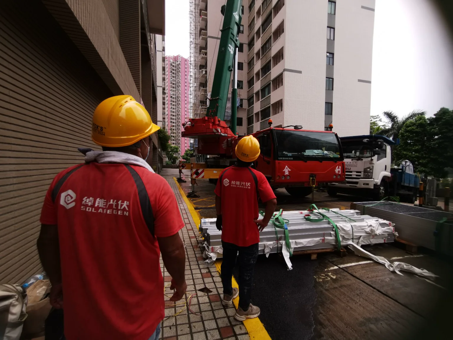 兩位身穿紅色綽能光伏工作服及安全帽的人員在吊車旁安排太陽能組件進場。